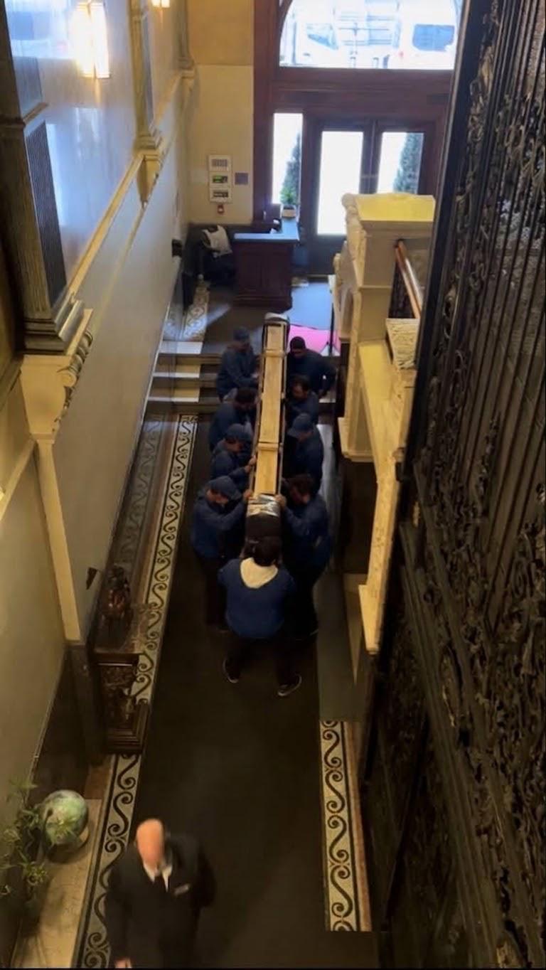 Une dizaine de déménageurs en uniforme transportant à la main une pièce de grande longueur dans le hall d'un édifice patrimonial